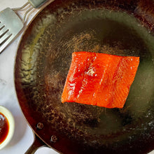 Salmon fillet cooking in a frying pan with a small bowl of sauce and a fork on a marble surface.