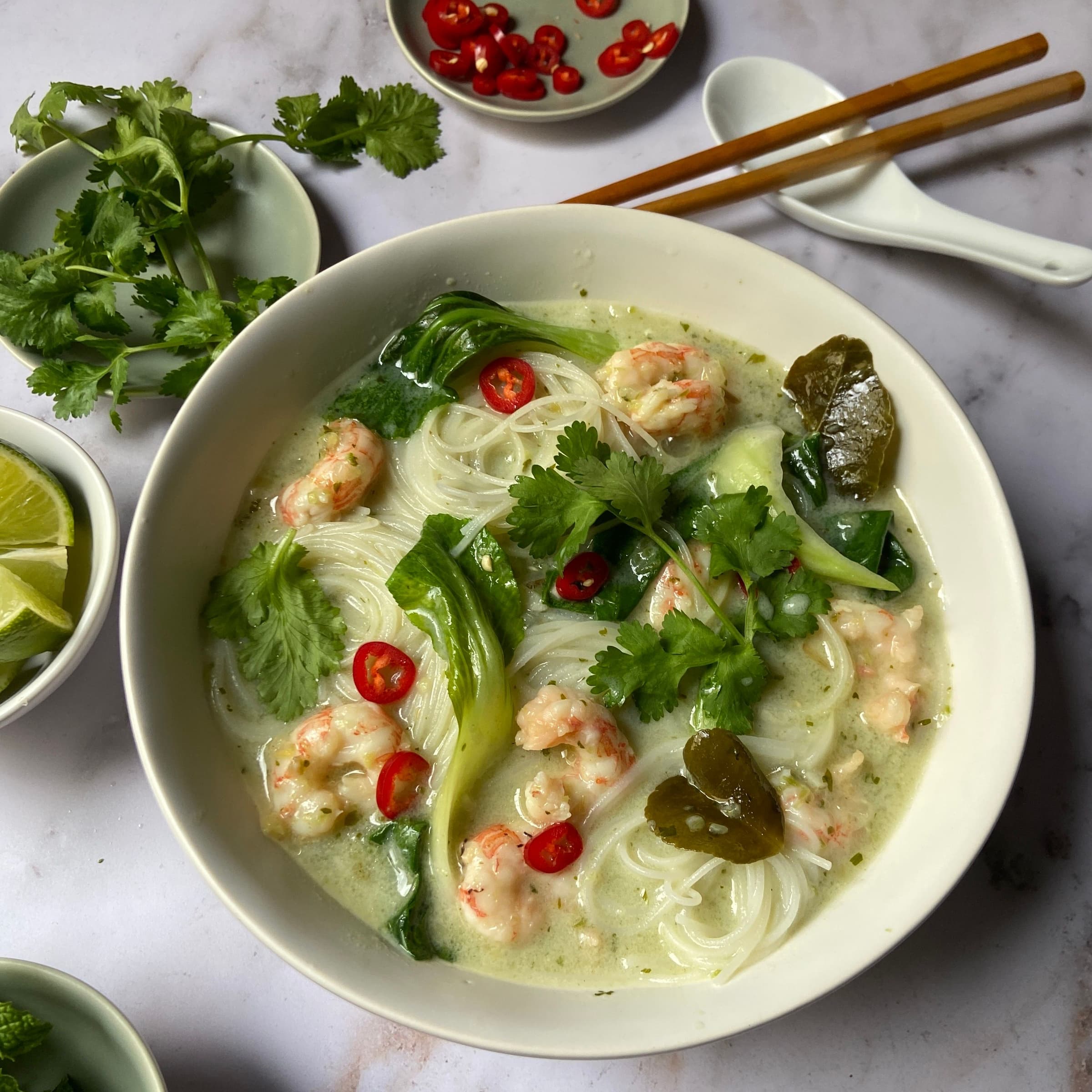 bowls of noodle soup, limes, herbs, and red chiles on a table