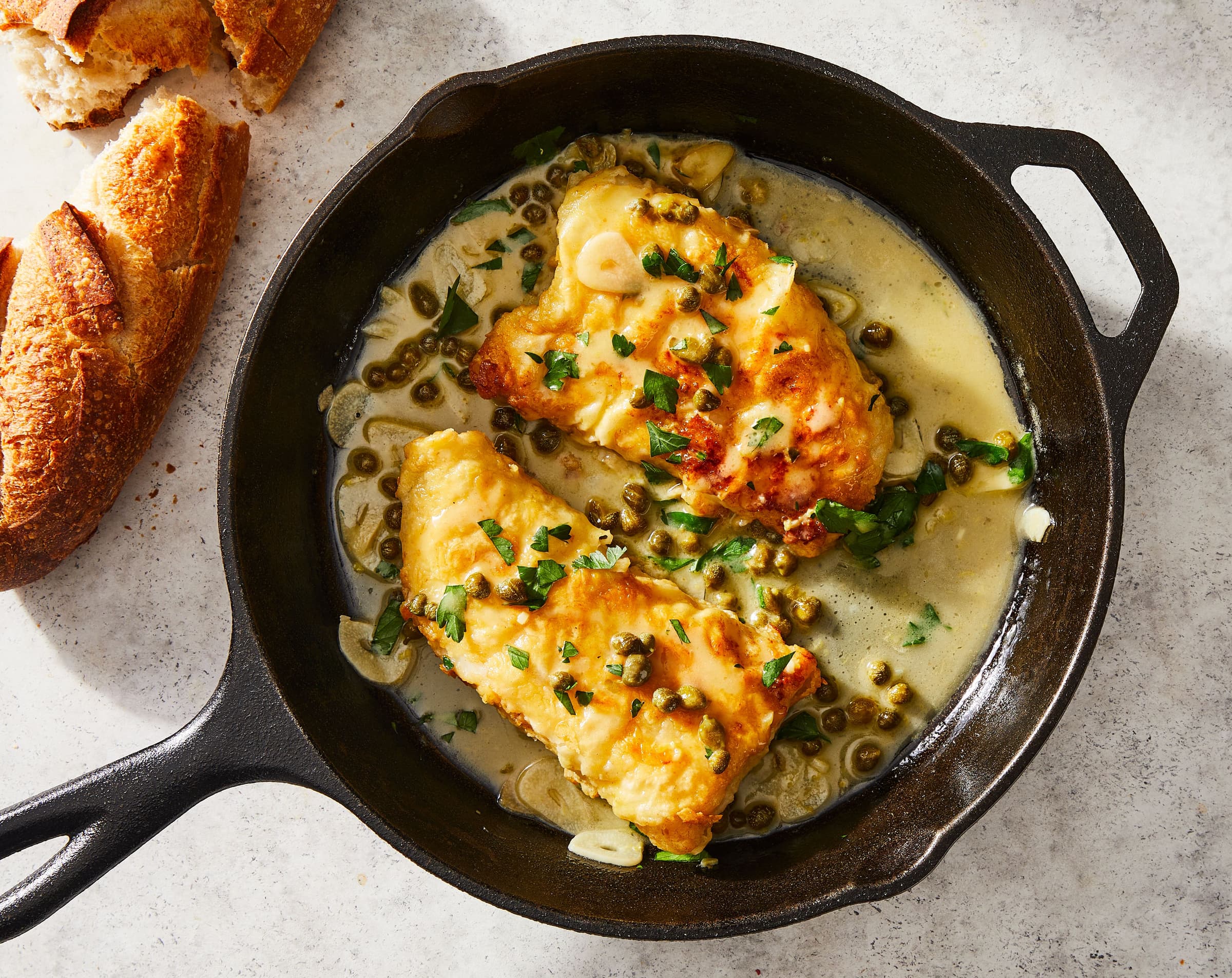 fish in a skillet with capers, herbs, and bread