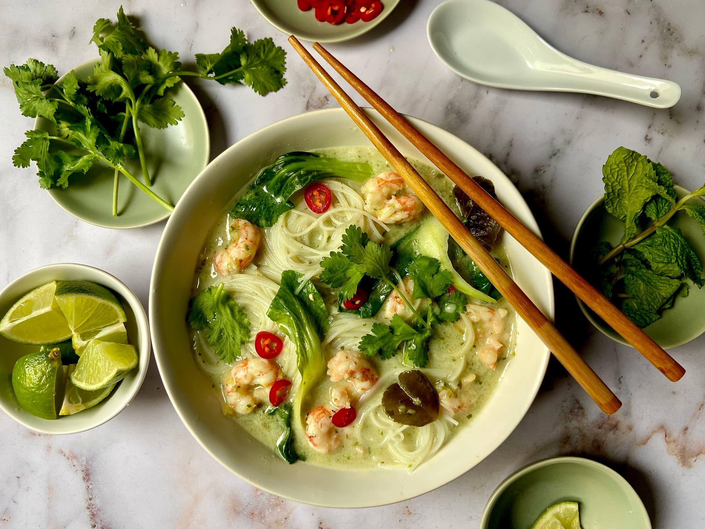 bowls of noodle soup, limes, herbs, and red chiles on a table