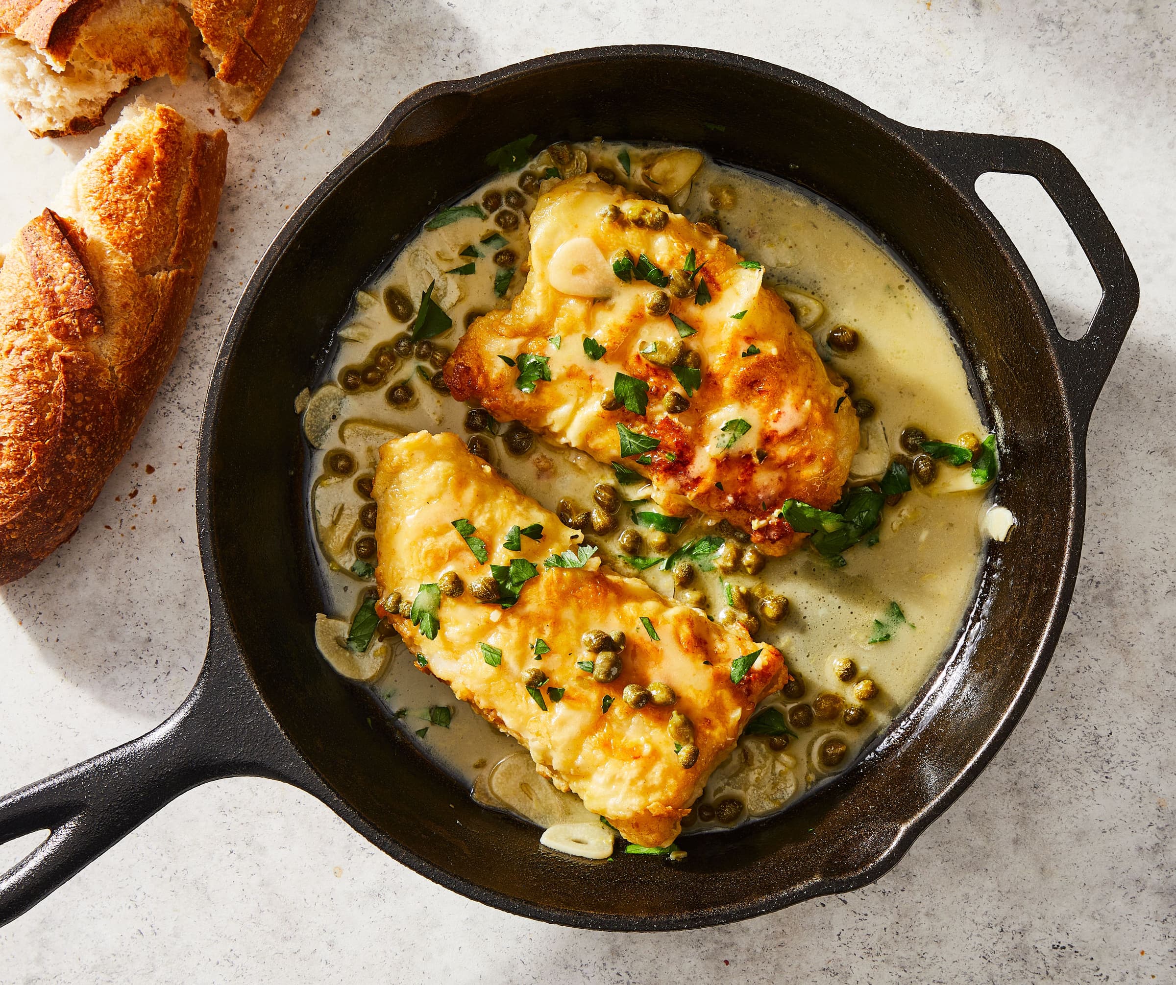 fish in a skillet with capers, herbs, and bread